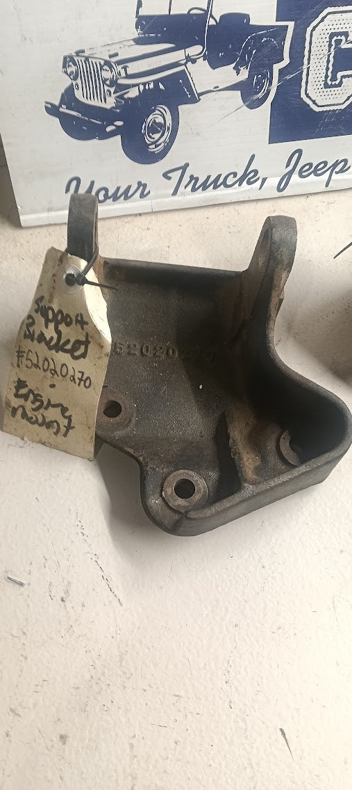 A used cast metal bracket, likely for a truck or jeep, with a handwritten paper tag attached. The background shows a white surface and a sign with a jeep illustration.