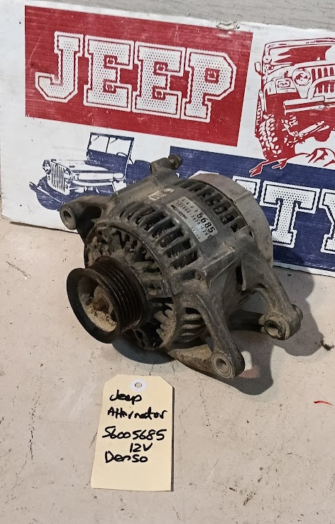 A used automotive alternator with a pulley and belt housing, photographed on a concrete floor in front of a painted Jeep-themed sign. A handwritten tag below the unit reads 'Jeep Alternator 56005685 12V Denso'.