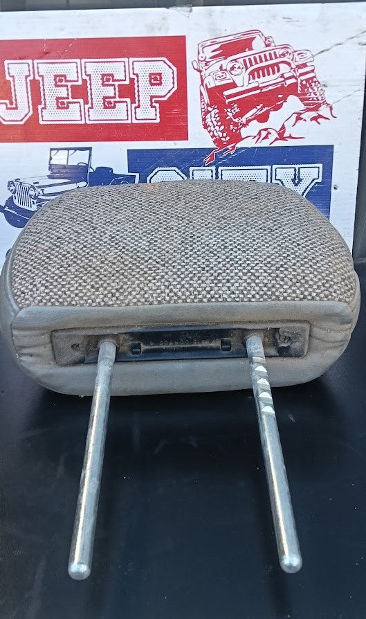 A used beige woven fabric headrest pillow with two metal mounting pins, shown against a background featuring a vintage red and white Jeep sign.