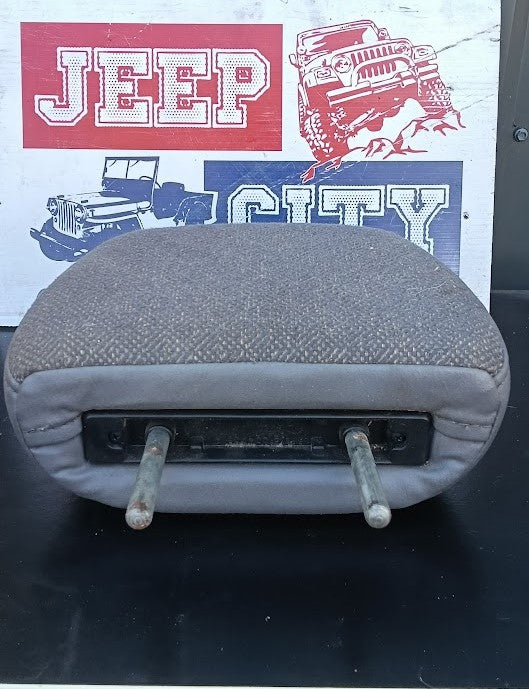 A used gray fabric car headrest or seat cushion with two metal support rods, shown against a background featuring a 'Jeep City' sign.